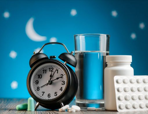Sobre uma mesa de cabeceira num quarto: Um despertador, um copo de água, uma cartela de cápsulas e um frasco de suplementos para dormir