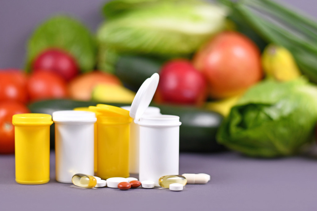 Potes de comprimidos de vitaminas e suplementos alimentares, incluindo cápsulas gelatinosas. Ao fundo, em segundo plano, estão frutas e verduras, simbolizando o que é princípio ativo